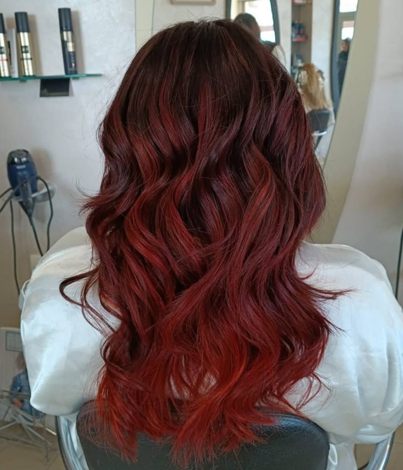 Capelli lunghi mossi sfumati dal bordeaux al rosso acceso su cliente di spalle con mantella bianca in un salone